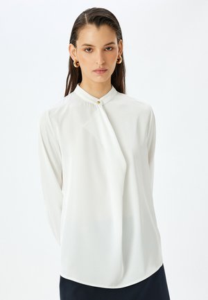 Blusa blanca de manga larga con cuello mao, que presenta un botón dorado y un diseño frontal drapeado. Tejido suave y ligero con un ligero brillo.