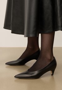 Svarta läderhögklackade skor med spetsig tådesign och en liten, utåtvinklad klack, som bärs med genomskinliga svarta strumpbyxor och en svart kjol.