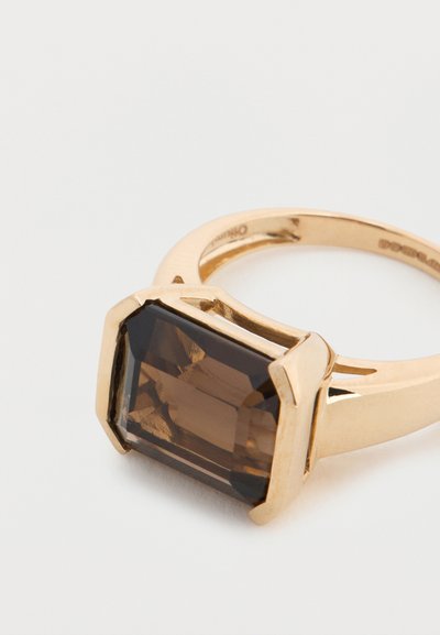 Δαχτυλίδι σε χρυσό τόνο με μεγάλο, ορθογώνιο καφέ πολύτιμο λίθο. Η μπάντα είναι λεία με γωνιώδεις λεπτομέρειες στο στήριγμα του λίθου.