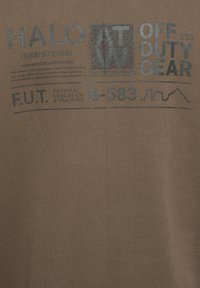 Brun bomullsskjorta med ett tryck i mörkgrå text, inklusive "HALO", "OFF DUTY GEAR" och "F.U.T." med en abstrakt linjedesign.