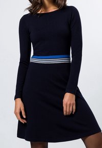 Marinblå stickad klänning med långa ärmar, med en horisontell blå och vit rand vid midjan. Slät textur, utfläkt silhuett.