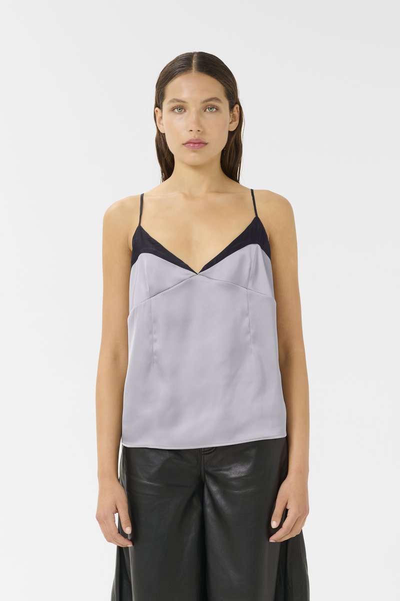 Camisola in satin grigia con spalline sottili, dettagli in navy lungo il scollo, scollatura a V e una texture liscia. Abbinata a pantaloni in pelle nera.
