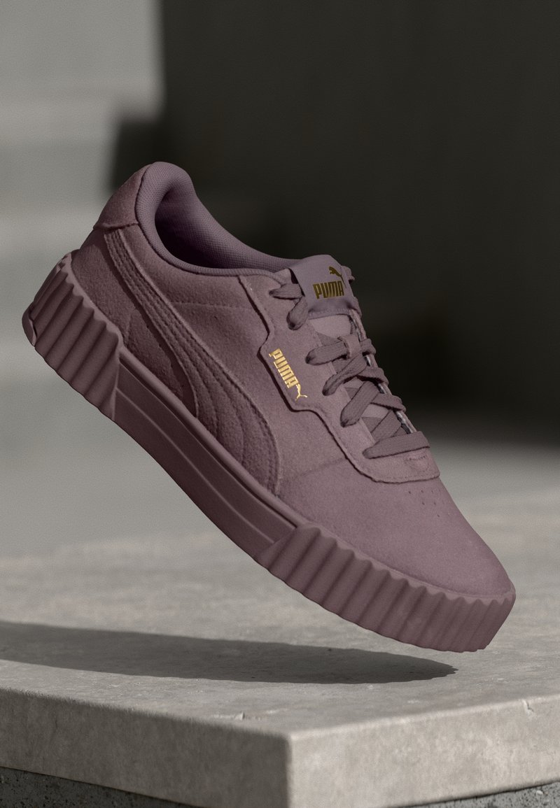 Baskets Puma violettes avec semelle épaisse rainurée, design à lacets et logo Puma doré sur la languette et le côté, suspendues au-dessus d'une surface en pierre.