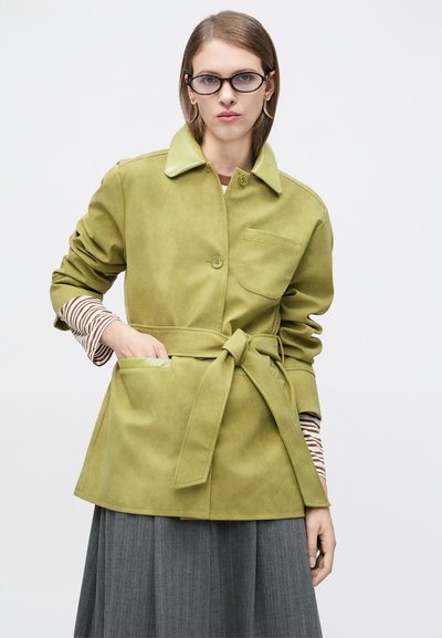 Γυναίκα που φοράει ανοιχτό πράσινο μπουφάν με ζώνη πάνω από ριγωτό πουκάμισο με μακριά μανίκια και γκρι πιετέ φούστα, με γυαλιά και χρυσά σκουλαρίκια.