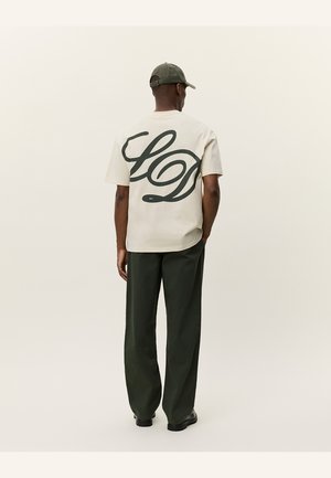 Beige short-sleeve shirt with large dark green cursive "LD" graphic on the back. Paired with dark green wide-leg pants and a matching cap.