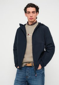 Μπλε ναυτικό μπουφάν με κουκούλα και εμπρόσθιο φερμουάρ, ελαφρύ ύφασμα και ελαστικές μανσέτες· φορεμένο πάνω από μπεζ φούτερ και μπλε τζιν.