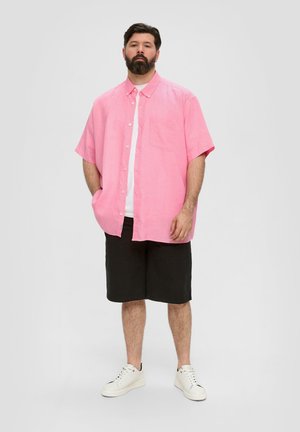 Kortärmige roze button-up shirt met een borstzak, gedragen over een wit T-shirt, gecombineerd met zwarte shorts en witte sneakers.