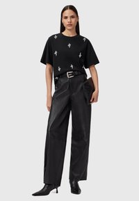 Kvinde iført en sort t-shirt med hvide slangeprints, sorte læder bukser med bred ben, sort bælte, som holder en sort clutch og sorte hæle støvler.