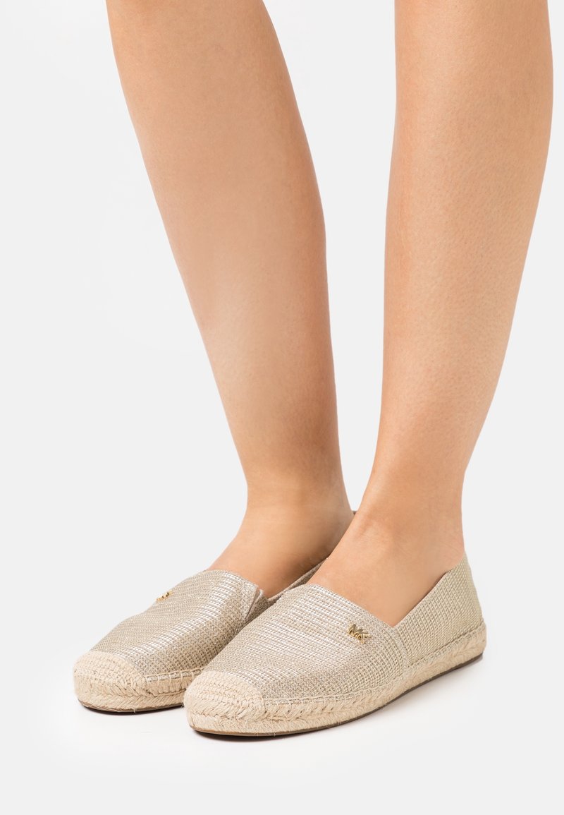 Jambes portant des espadrilles beige tissées à enfiler avec des semelles en jute tressé et un petit détail de logo doré sur un fond clair.