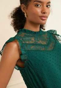 Grön klänning med spetsdetaljer, volangkrage, texturerat tyg, prickmönster och spetsaccenter på axlarna. Guldsmycken lyfter utseendet.