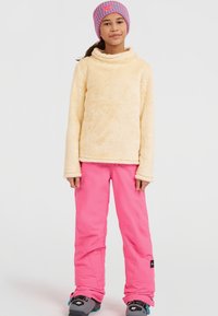 Fleece-Pullover in hellgelb mit hohem Kragen; kombiniert mit rosa Skihosen und grauen Schneestiefeln mit blauen Akzenten. Gemusterte Strickmütze.