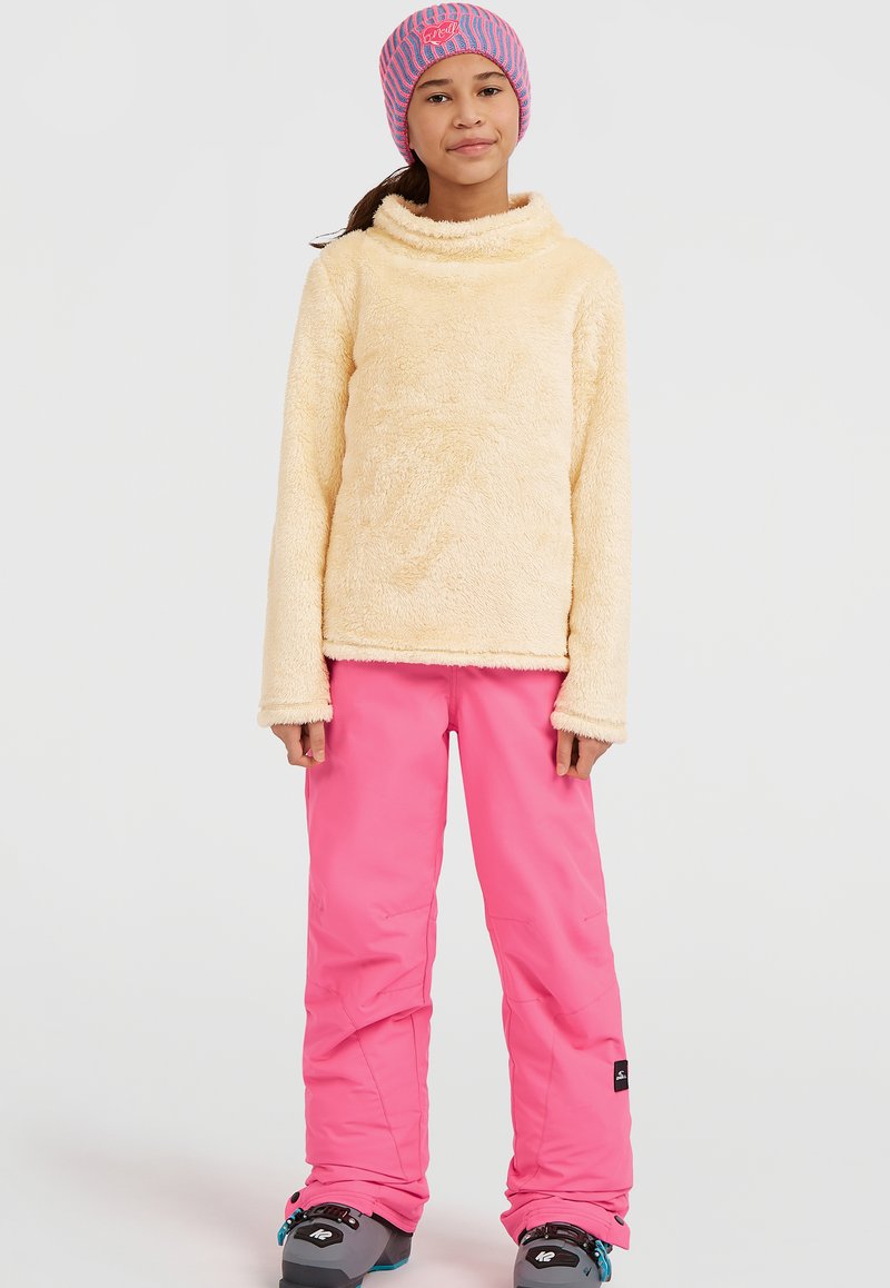 Fleece-Pullover in hellgelb mit hohem Kragen; kombiniert mit rosa Skihosen und grauen Schneestiefeln mit blauen Akzenten. Gemusterte Strickmütze.