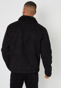 Mand med kort hår iført en sort jakke med shearling-krave og sorte jeans, vendt væk fra en ensfarvet lys baggrund.