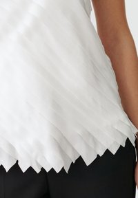 Top alb cu plişuri, cu un tiv ondulat, având benzi orizontale şi o textură netedă, asortat cu pantaloni negri pentru contrast.