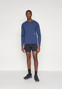 Ein Mann steht vor einem einfarbigen Hintergrund und trägt ein blaues Langarmhemd, schwarze Sportshorts, schwarze Socken und schwarze Laufschuhe.