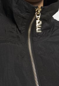 Svart jacka med hög krage, med en guldfärgad dragkedja och textilstruktur. Anmärkningsvärda detaljer inkluderar en glänsande, metallisk dragkedja.