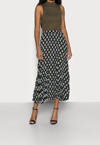 Olivgrön ärmlös topp i kombination med en midi kjol med svart och vit diamantmönster, som har en flowig design och en åtsittande midja. Svarta remsandaler.