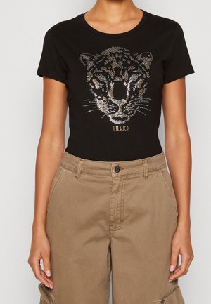 Zbliżenie osoby noszącej czarną koszulkę z wzorem twarzy lamparta z cyrkoniami oraz beżowe spodnie z wysokim stanem.