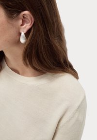 Orecchini a goccia bianchi realizzati in un materiale liscio, indossati con un maglione lavorato a maglia di un beige chiaro. I capelli sono marroni e leggermente ondulati.