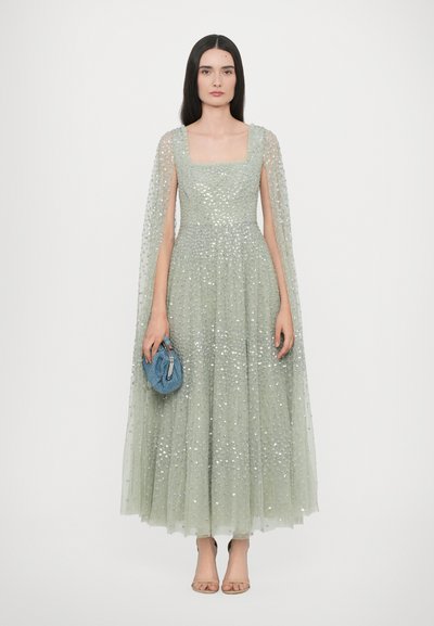 Lichtgroene avondjurk met een getailleerde top, paillettenversieringen en een vloeiende cape. Houdt een kleine blauwe handtas vast, met open teed schoenen.