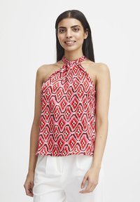Top a halter rossa e rosa con motivo geometrico su un tessuto morbido, abbinato a pantaloni bianchi.