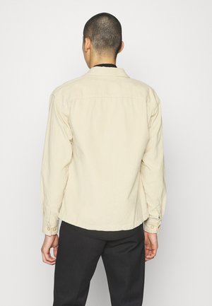 Mann mit kurzen Haaren, der ein beiges Langarmhemd und schwarze Hosen trägt und mit dem Rücken zu einer schlichten Hintergrund steht.