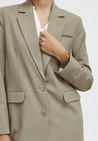 Světle hnědý blazer s dvěma knoflíky, klopovými límci a jednou kapsou na hrudi. Tkanina má jemnou strukturu a ploché přední kapsy.