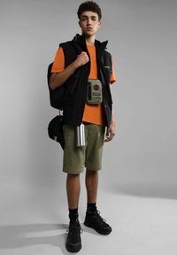 Gilet nero sopra una t-shirt arancione, pantaloni cargo verdi, sneakers nere. Gli accessori includono una pochette verde e una bottiglia d'acqua argentata.
