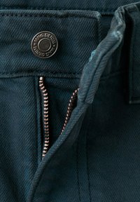 Gros plan sur un jean en denim bleu foncé avec une fermeture éclair partiellement ouverte et un bouton en métal marqué "MNG DENIM".