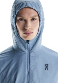 Veste à capuche bleu clair avec une texture lisse, fermeture éclair intégrale et un logo noir sur la poitrine. La capuche est bien ajustée.