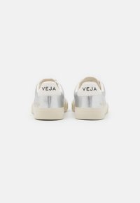Silver metallic sneakers with cream accents, featuring a textured surface, flat beige sole, and "VEJA" branding on the heel and sides.