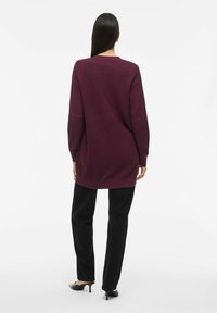 Langer, übergroßer Strickpullover in Dunkelpink, mit strukturiertem Stoff und seitlichen Schlitzen. Kombiniert mit schwarzen Hosen und eleganten schwarzen Absätzen.