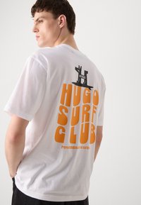 T-shirt bianca in cotone con grafica arancione "HUGO SURF CLUB" e logo nero sul retro. Maniche corte, vestibilità comoda e texture morbida.