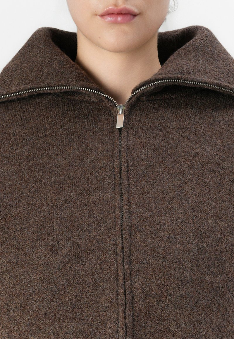 Bruine wollen zip-up jas met een hoge kraag, voorzien van een zilveren rits en een gestructureerde stof, die warmte en een gezellige uitstraling benadrukt.