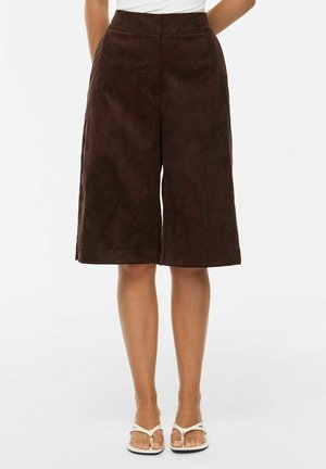 Shorts marron large jusqu'au genou, portés avec des sandales blanches et un haut blanc, modèle debout sur un fond uni.