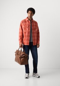 Camisa de cuadros rojos con dos bolsillos en el pecho, camiseta gris por debajo, jeans oscuros y bolso de cuero marrón, complementado con zapatillas deportivas plateadas.
