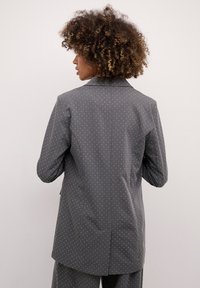 Pelēks žakets ar baltiem punktiņiem, piegriezts siluets, viens aizmugurējais vents un asas atlokas. Auduma tekstūra izskatās gluda un strukturēta.