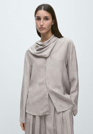 Kvinde med langt brunt hår iført løs, lysegrå draperet bluse og matchende plisseret nederdel foran en ensfarvet lys baggrund.