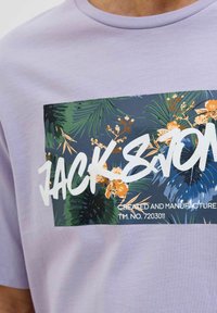 Fialové tričko s krátkým rukávem, které má velký obdélníkový potisk se tropickým listím a výrazným bílým logo "JACK&J".