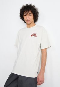 Svetlo béžové bavlnené tričko s krátkymi rukávmi, na ľavej hrudi s červeným logom Nike SB. Jednoduchý, voľný strih.