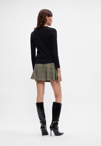 Fekete hosszú ujjú felső, zöld kockás mini szoknyával és térdmagasságú fekete fényes sarkú csizmával párosítva. Sima anyag és strukturált kialakítás.