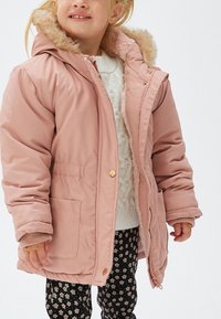 Niño con un abrigo de invierno rosa claro con capucha y borde de piel, suéter blanco de punto y pantalones negros con patrón de flores blancas.