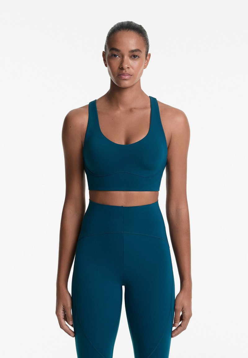 Soutien-gorge de sport teal avec de larges bretelles et un col arrondi, associé à des leggings teal taille haute présentant des détails de couture courbés.