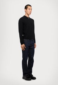 Maglione nero a maglia con colletto, abbinato a jeans in denim blu scuro e scarpe nere. Il modello sta in piedi, con le mani rilassate sui fianchi.
