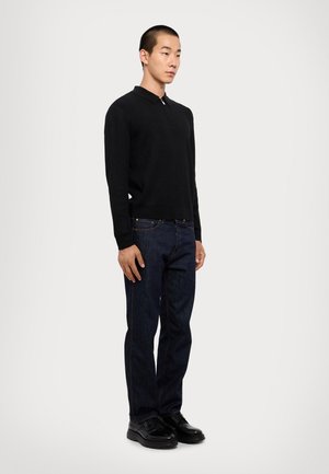 Maglione nero a maglia con colletto, abbinato a jeans in denim blu scuro e scarpe nere. Il modello sta in piedi, con le mani rilassate sui fianchi.
