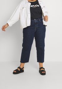 Persoon gekleed in donkerblauwe cropped jeans, zwart Levi's shirt, witte jas en zwarte open-tee sandalen, staand tegen een effen achtergrond.