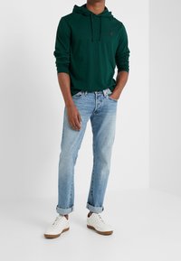 Grön långärmad hoodie med snörning, ljus tvättade tapered jeans och vita sneakers med beige sulor. Avslappnad ensemble, minimal mönster.