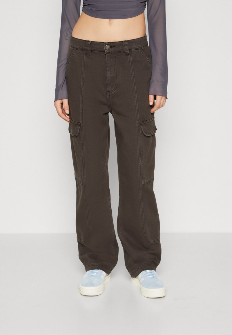Pantalons cargo taille haute en coton marron foncé, avec des poches latérales et des coutures contrastées. Portés avec un haut transparent à manches longues et des baskets bleu clair.