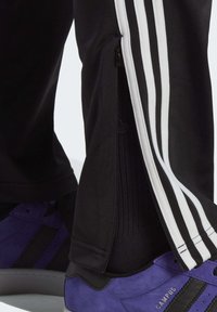 Pantalones deportivos negros con rayas blancas, con aperturas con cremallera en los tobillos. Combinados con zapatillas de ante moradas.