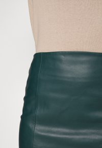 Jupe en cuir verte à texture lisse et coupe près du corps. La ceinture est mise en valeur par un haut tricoté beige.